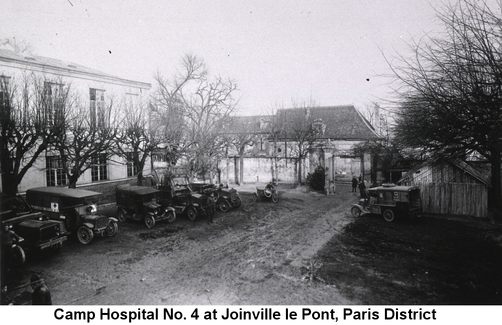 Black and white photo of several large white buildings; on the left a two-story building with large vertical windows is fronted by a line of leafless trees, before which stand several parked ambulances in a muddy parking lot. In the distance a large single-story building with a high steep roof is partially obscured by more leafless trees.